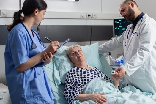 Doctor checking senior patient in hospital intensive care, checking oxygen saturation, blood pressure on oxymeter attached on sick senior patient while nurse making notes on clipboard for diagnosis.