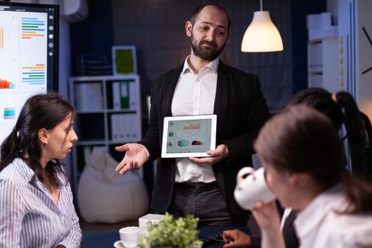 Workaholic overworked businessman explaining company strategy using tablet for presentation working overtime in meeting office room late at night. Diverse multi-ethnic teamwork brainstorming ideas.