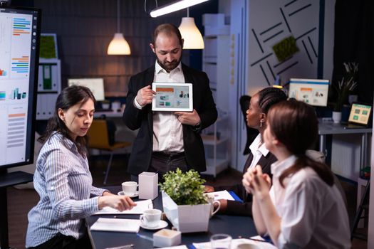 Focused businessman showing corporate graphs presentation using tablet working at company ideas. Diverse multi-ethnic businesspeople brainstorming strategy late at night in office meeting room