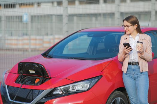 Girl Use Coffee Drink While Using Smart Phone and Waiting Power Supply Connect to Electric Vehicles for Charging the Battery in Car.
