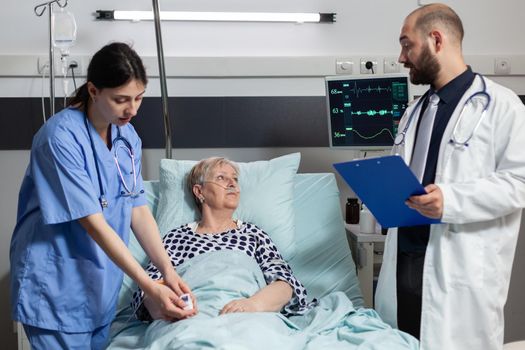 Medical nurse attaching oxymeter on senior woman patient, laying on hospital bed discussing with doctor about diagnosis and treatment while she breaths with help from oxygem mask.