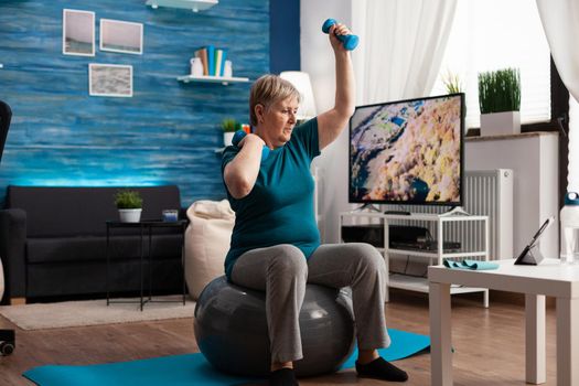 Focused retirement pensioner sitting on fitness swiss ball raising hand streching arm muscle doing healthcare exercise. Senior woman exercising body resistance using dumbbells in living room