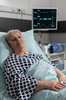 Senior woman patient laying in hospital bed following recovery treatment, getting receiving a medication through an intravenous line. Oxymeter attached on finger monitoring oxygen blood saturation.