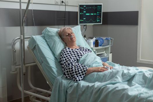 Senior woman resting in hospital bed breathing with help from oxygen mask, because of pulmonary illness. Getting intravenous medicine from iv drip bag during recovery therapy.