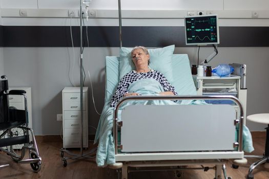 Senior woman patient laying in hospital bed with oxymeter attached on body, measuring oxygen saturation from blood and blood pressure and bmp. Getting treatment from iv drip bag.