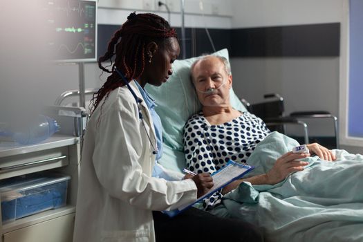 African american doctor in hospital room discussing diagnosis and treatment with sick senior man laying in bed reassuring full recovery. Unwell patient breathing through oxygen mask.