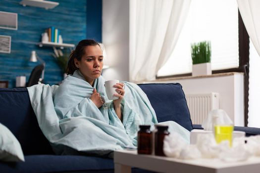 Woman with cold and flu drinking medicine for infection and fever wrapped in blanket. Seasonal pain headache symptoms sitting on sofa tired, taking sickness disease treatment