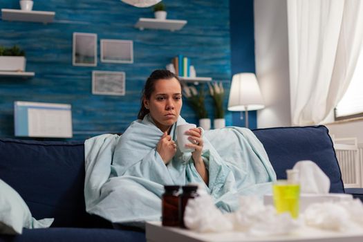 Woman with cold and flu drinking medicine for infection and fever wrapped in blanket. Seasonal pain headache symptoms sitting on sofa tired, taking sickness disease treatment