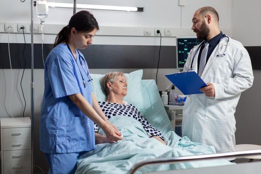 Nurse reading blood oxygen saturation from oxymeter device attached on sick senior woman patient laying in hospital bed resting. Doctor monitoring patient health condition.