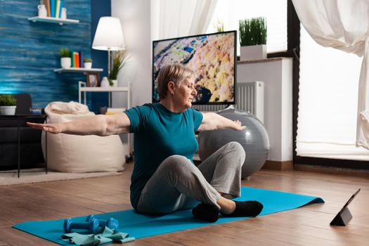 Retired senior woman searching fitness tutorial on laptop sitting on yoga mat streching arm during wellness workout in living room. Pensioner training body muscles working at flexibility