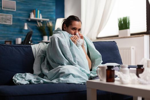Woman with sickness wrapped in blanket at home ill feeling unwell. Young caucasian adult suffering from temperature fever disease illness flu, taking medical treatment sitting on couch