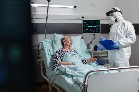 Senior woman patient laying unconscious in hospital bed during coronavirus outbreak, breathing with help from oxygen mask. Doctor dressed in ppe suit to prevent infection.