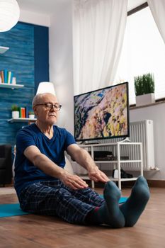 Retirement senior man sitting on yoga mat stretching legs muscles during body training working at flexibility. Pensioner in sportswear slimming weight during muscle resistance in living room