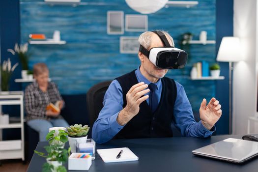Happy pensioner experiencing virtual reality wearing headset. Elderly man pensioner in living room sitting at desk with futuristic goggles for augmented cyberspace wife watching tv.