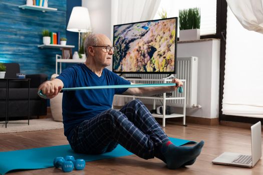 Retirement senior man sitting on yoga mat with leg in crossed position stretching arms muscles using stretch elastic band during sport routine in living room. Pensioner exercising bodyresistance