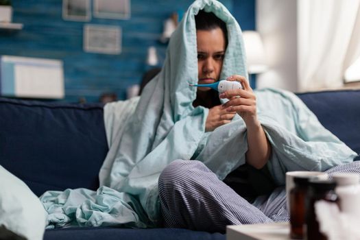 Caucasian woman measuring temperature ill at home with a thermometer. Person feeling sick cold unwell, checking fever and symptoms of disease flu infection. Resting adult with headache