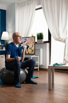 Senior man training body resistance doing arms exercise sitting on swiss ball in living room. Pensioner watching online wellness training on laptop exercising muscle workout using dumbbell
