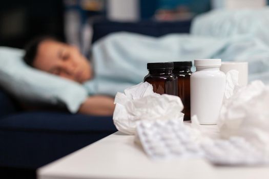 Close up of medication, pills and napkins for sick woman sleeping on sofa. Taking drugs and medical treatment against cold flu virus symptoms. Young adult with infection and temperature.