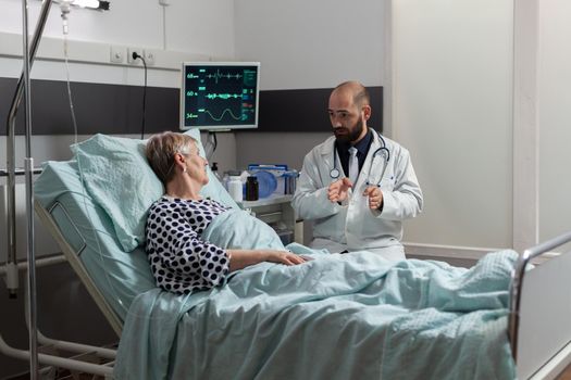 Doctor sitting next so sick unwell senior patient laying in bed during friendly discussion about treatment. Breathing with help from oxygen tube and getting medication through iv drip bag.