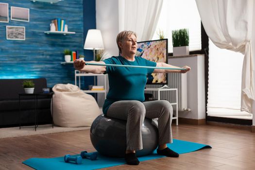 Retired pensioner sitting on swiss ball working at body weight stretching muscle arm using fitness elastic band. Active senior woman watching online sport video on laptop practicing strech exercise