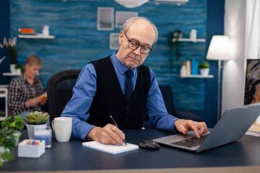 Senior businessman writing notes wearing glasses working in home office. Elderly man entrepreneur in home workplace using portable computer sitting at desk while wife is reading a book sitting on sofa.