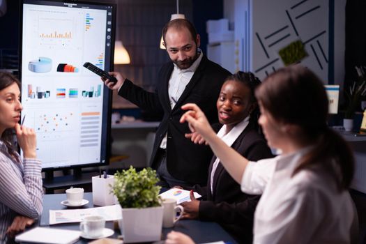 Entrepreneur man brainstorming management strategy working hard in meeting office room late at night. Diverse multi-ethnic business team looking at financial company presentation on monitor.