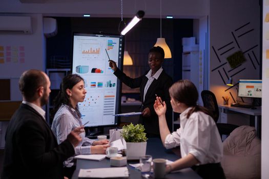 Overworked businesswoman with dark skin brainstorming financial strategy overworking in company meeting room late at night. Workaholics multi-ethnic teamwork discussing marketing deadline project.