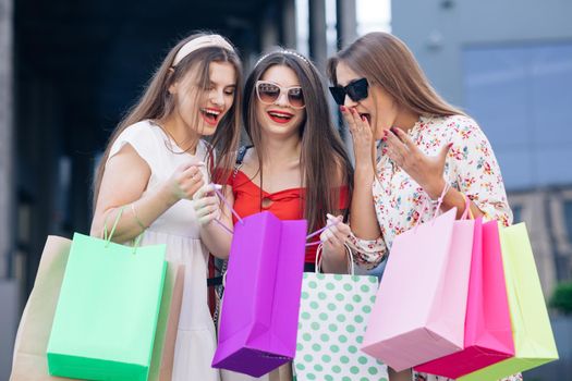 Successful shopping. Casual makeup. A group of young happy cute woman in casual dresses, top and pants walking from the building with yellow, green, purple and pink bags in their hands.