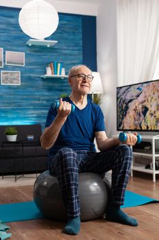 Retirement senior man sitting on swiss ball exercising arms muscles doing fitness exercises using workout dumbbells. Focused pensioner training body strength resistance in living room