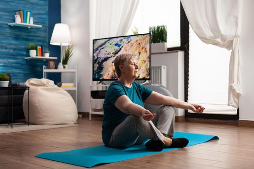 Peaceful retired senior woman meditate during meditation retreat workout sitting comfortable in lotus position with closed eyes on yoga mat. Caucasian female practicing body exercise in living room
