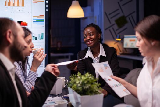 Workaholics focused multi-ethnic businesspeople overworking in company office meeting room brainstorming ideas late at night. Coworkers sitting at desk analyzing statistics paperwork