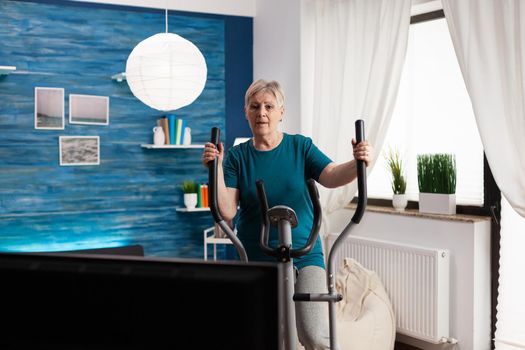Focused senior woman doing workout training body muscle watching online video gym exercise on television using slimming bicycle at home in living room. Fitness pensioner working at cardio resistance