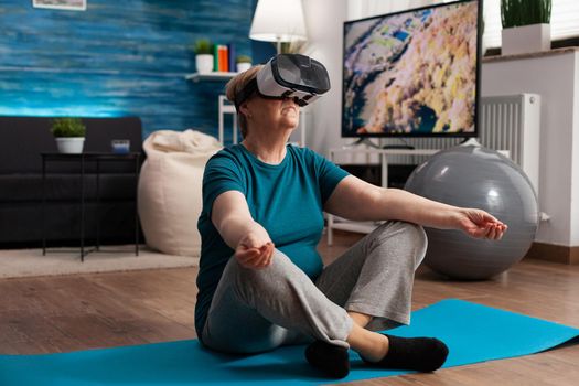 Healthy senior woman using virtual reality headset meditating while sitting in lotus position on yoga mat in living room during meditation workout. Pensioner training body muscle doing sports exercise