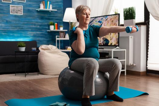Athletic pensioner in sportswear watching online aerobic workout using tablet sitting on swiss ball exercising body resistance. Senior woman doing arms exercise using dumbbells in living room
