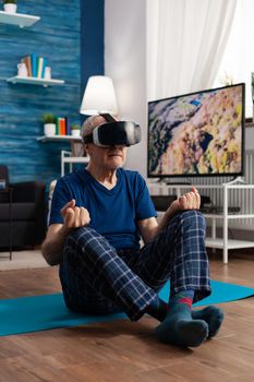 Senior man wearing virtual reality headset practicing meditation exercise sitting on yoga mat in living room stretching body muscle. Pensioner meditate during healthy workout in lotus position