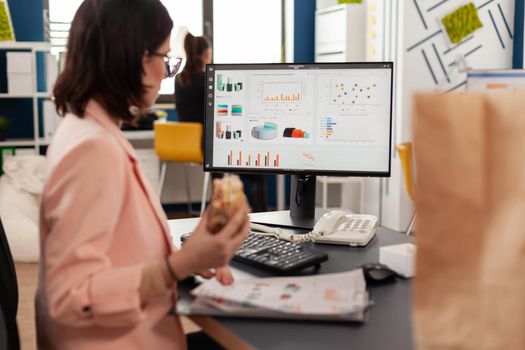 Businesswoman eating tasty sandwich having meal break working in business company office during takeout lunchbreak. Fast food order paper bag delivered at workplace. Woman typing strategy on computer.