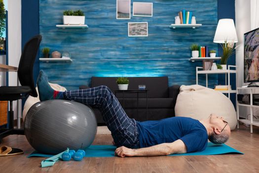 Retired senior sportman sitting on yoga mat practicing warming legs up exercises using swiss ball stretching abdominals muscles. Focused pensioner training body strength resistance in living room