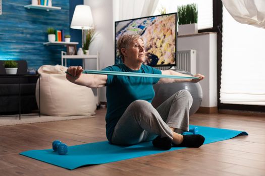Retired senior woman sitting on yoga mat in lotus position stretching arms muscles using stretch elastic band during wellness sport routine in living room. Pensioner exercising body resistance