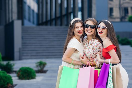 Girls after shopping.Three women hold in hands colorful shopping bags. Consumerism, purchases, sales, lifestyle.