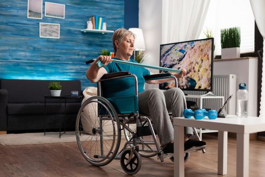 Invalid senior woman in wheelchair holding resistance elastic band stretching body muscle recovering after disability accident watching workout video on tablet. Pensioner doing aerobics exercise