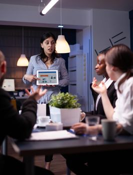 Business leader in meating room late at night discussing with her team holding tablet pc with charts. Diverse multi-ethnic teamwork solving financial strategy. Team working on deadline.