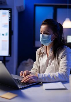 Project manager with protection face mask using professional laptop for public marketing late at night in business office during global pandemic. Tired woman working with modern technology network