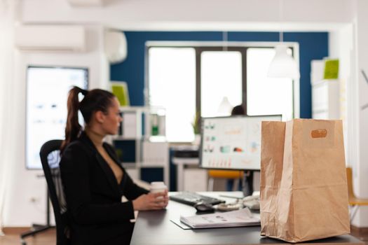 Close up shot of paper bag with tasty delicious meal takeaway food, of business woman working on computer analyzing company documents, graphs. Employee holding coffee cup.