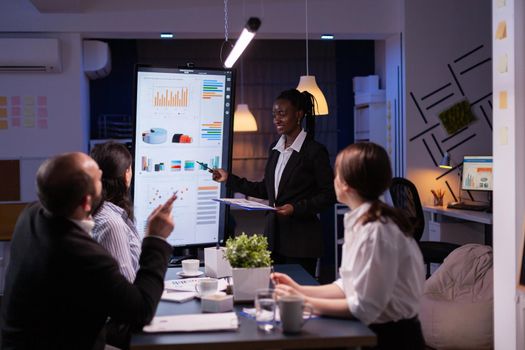 Workaholic african american leader standing in front of presentation monitor explaining marketing project solution late at night in company meeting room. Diverse teamwork working at business ideas.