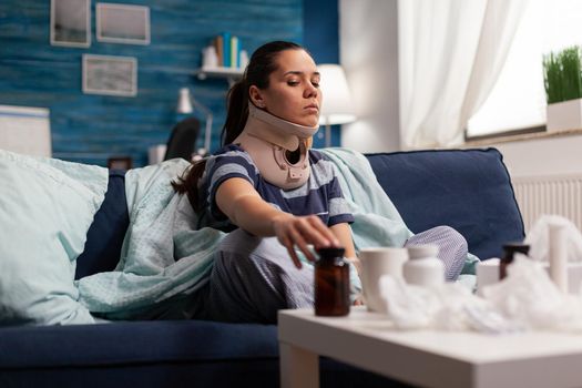 Woman in neck brace suffering from pain on sofa after bad accident injury. Young caucasian adult with ache taking medicine for back complication, physical muscular contracture