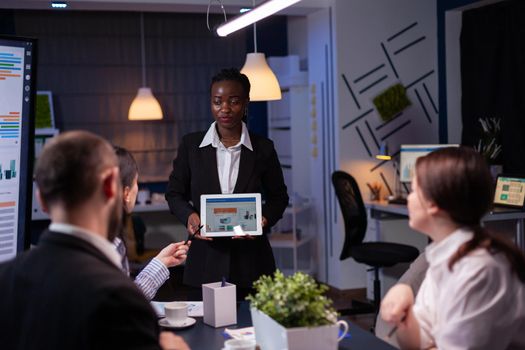 Focused workaholic entrepreneur woman with dark skin explaining management strategy using tablet. Business diverse multi-ethnic teamwork overworking in company office meeting room late at night