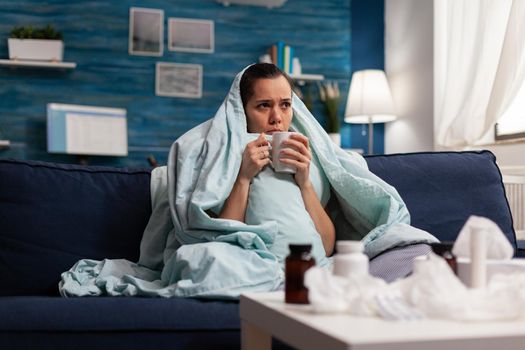 Sick woman sitting at home in blanket with hot tea suffering from fever temperature infection flu headache cold. Young person drinking tea, with seasonal virus symptoms feeling unwell