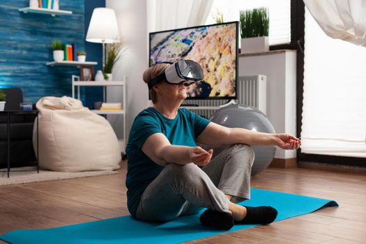 Retreat senior woman sitting on yoga mat in lotus position in living room wearing virtual reality headset during pilates meditation workout. Pensioner stretching body muscle doing training exercise