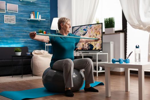 Cheerful retired pensioner working arm muscles using elastic band practicing aerobic exercise. Pensioner sitting on swiss ball in living room working at body healthcare resistance watching gym video