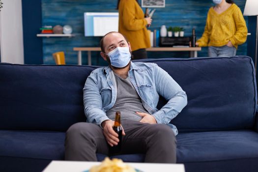 Man enoying spending time with friends in living room keeping social distancing wearing face mask to prevent corovnavirus spreading in the course of global outbreak holding beer bottle.